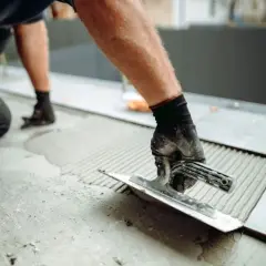 Układanie płytek 60x60 wymaga odpowiedniego kleju. Ręka w rękawicy rozprowadza zaprawę kielnią na podłożu.