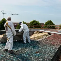 Dwóch pracowników w białych kombinezonach usuwa eternit z dachu. Jeden z nich trzyma duży, pofalowany arkusz.