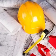 Plany budowy domu systemem gospodarczym: żółty kask, miarka, poziomica, ołówki i czerwone kształty domów na tle rysunków technicznych.