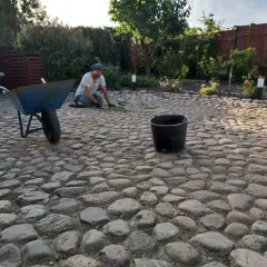 Mężczyzna pracuje przy budowie podjazdu z kamienia polnego. Obok stoi taczka i wiadro.