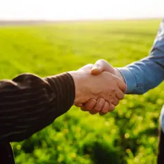 Dwaj rolnicy uścisnęli sobie dłonie na tle zielonego pola. Umowa dotycząca podziału działki rolnej na mniej niż 30 arów została zawarta.