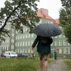 Osoba z parasolem idzie chodnikiem w deszczu. W tle zielony budynek i drzewa. Czyżby to był podatek od deszczu?