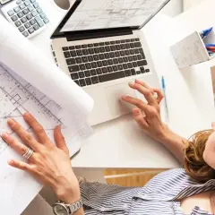 Kobieta analizuje plan zabudowy lub szkic sytuacyjny, pracując przy laptopie.