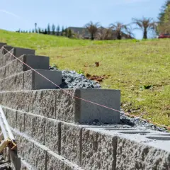Budowa muru oporowego z bloczków betonowych. Widać drabinę i różową nić, która pomaga w równym układaniu.