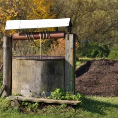 Stary, betonowy krąg studni kręgowej z drewnianą konstrukcją i metalową korbą. W tle jesienne drzewa i kopiec ziemi.