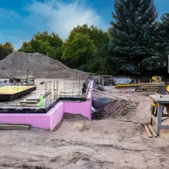 Budowa w stanie zero: fundamenty z różową izolacją, zbrojenie, sterta piasku i sprzęt budowlany.