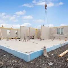 Budowa domu z prefabrykatów. Na placu budowy widać fundamenty, ściany z oknami i instalacjami, a nad nimi uniesiony element przez dźwig. Czy to keramzyt?