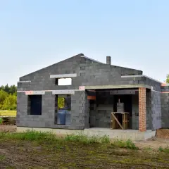 Budowa domu z keramzytu w otoczeniu zieleni. Ściany z szarego pustaka, widoczne otwory okienne i drzwiowe.