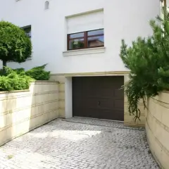 Podjazd z kostki prowadzi do brązowej bramy garażowej. Otaczają go kamienne murki z bujną zielenią.
