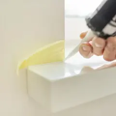 Ręka z pistoletem do silikonu aplikuje uszczelniacz w narożniku, zabezpieczonym taśmą. Oto jak położyć silikon w narożnikach.