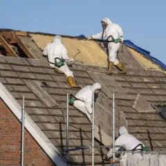 Pracownicy w kombinezonach ochronnych usuwają stare pokrycie dachowe. To praca z materiałami zawierającymi azbest, co to jest, jest niebezpieczne.