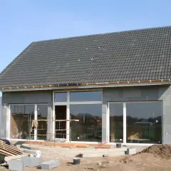 Dom w stanie surowym zamkniętym, z dachem pokrytym dachówką i dużymi oknami. Wokół plac budowy z materiałami.
