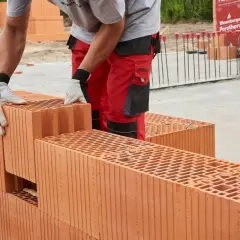 Budowa domu z pustaków ceramicznych Porotherm. Pracownik w czerwonych spodniach i rękawicach układa kolejne elementy.