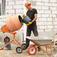 Budowlaniec miesza beton B20, odmierzając składniki do betoniarki. Obok stoi taczka i łopata.