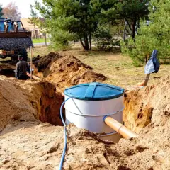 Trwa montaż oczyszczalni przydomowej. Koparka zasypuje wykop, a pracownicy nadzorują instalację zbiornika.
