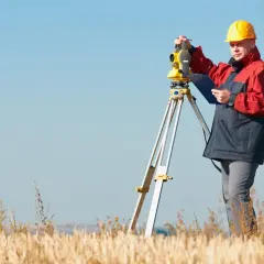 Geodeta w żółtym kasku i czerwonej kurtce pracuje z tachimetrem w polu. Zastanawiasz się, ile bierze geodeta za wytyczenie działki?