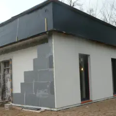 Budowa domu ze styropianu w toku. Ściany z bloczków, częściowo otynkowane, dach przykryty czarną folią.