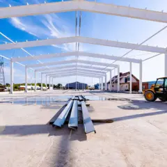Widok na budowę hali stalowej z betonową posadzką, na której stoją stalowe elementy konstrukcyjne. W tle żółty ładowarka teleskopowa.
