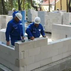 Budowa domu z bloczków. Pracownicy w niebieskich kombinezonach układają ściany. Wybór materiału: solbet czy ytong?