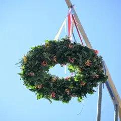 Wieniec z zieleni i kwiatów, ozdobiony wstążkami, wisi na drewnianym słupie. To tradycyjna wiecha na dach, symbolizująca pomyślność.