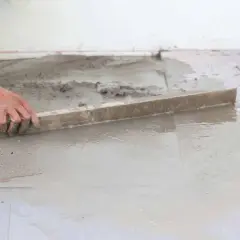 Ręka wyrównuje świeżą posadzkę na gruncie za pomocą łaty.