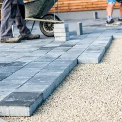Układanie płyt betonowych na podjeździe – alternatywa dla kostki. Pracownicy budowlani układają szare płyty na podsypce żwirowej.
