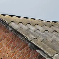 Stary dach pokryty eternitem, czyli materiałem zawierającym azbest, przy ceglanej ścianie.