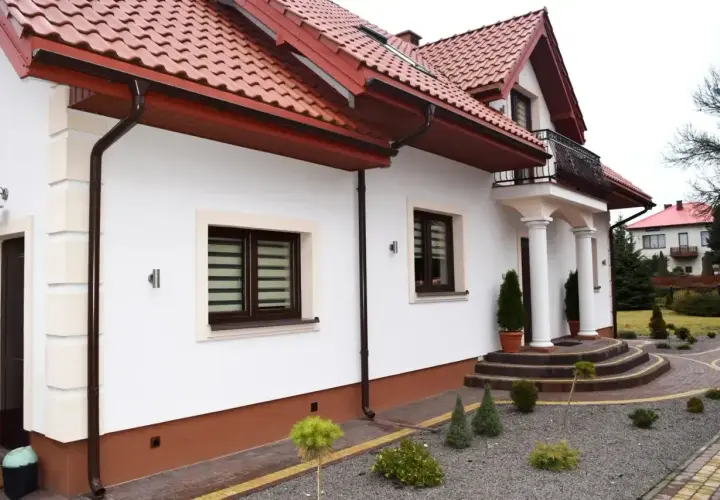 Biały dom z brązowym dachem i cokołem. Eleganckie schody prowadzą do wejścia.