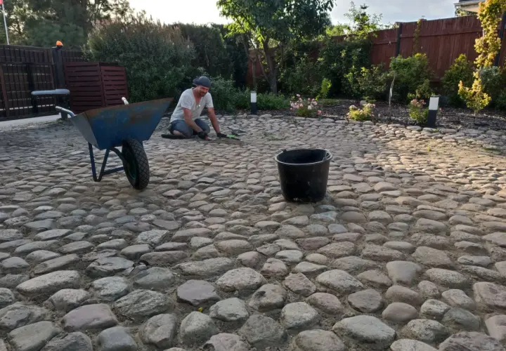 Mężczyzna pracuje przy budowie podjazdu z kamienia polnego. Obok stoi taczka i wiadro.