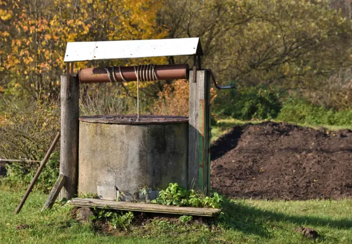 Stary, betonowy krąg studni kręgowej z drewnianą konstrukcją i metalową korbą. W tle jesienne drzewa i kopiec ziemi.