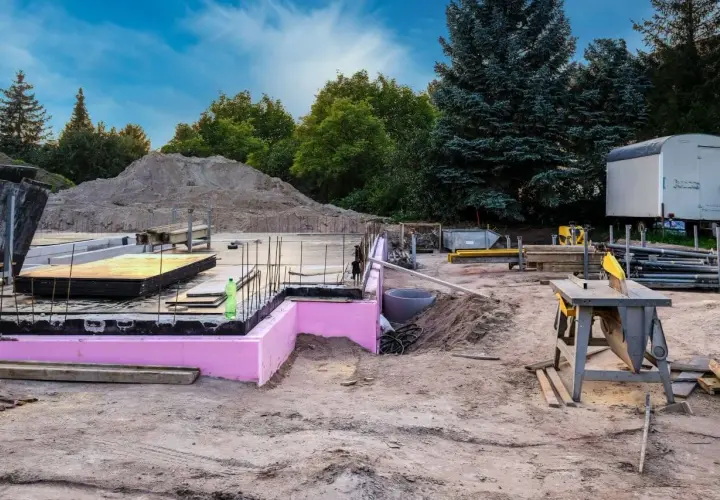 Budowa w stanie zero: fundamenty z różową izolacją, zbrojenie, sterta piasku i sprzęt budowlany.