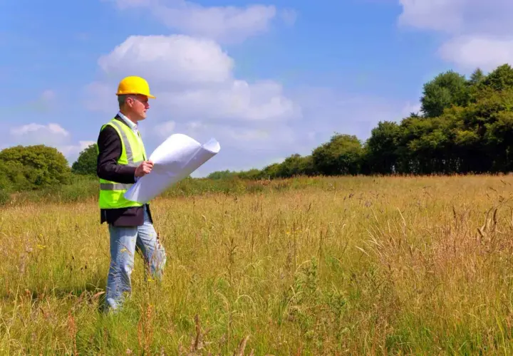 Inżynier w kasku i kamizelce odblaskowej analizuje plany podczas badań geotechnicznych gruntu na łące.