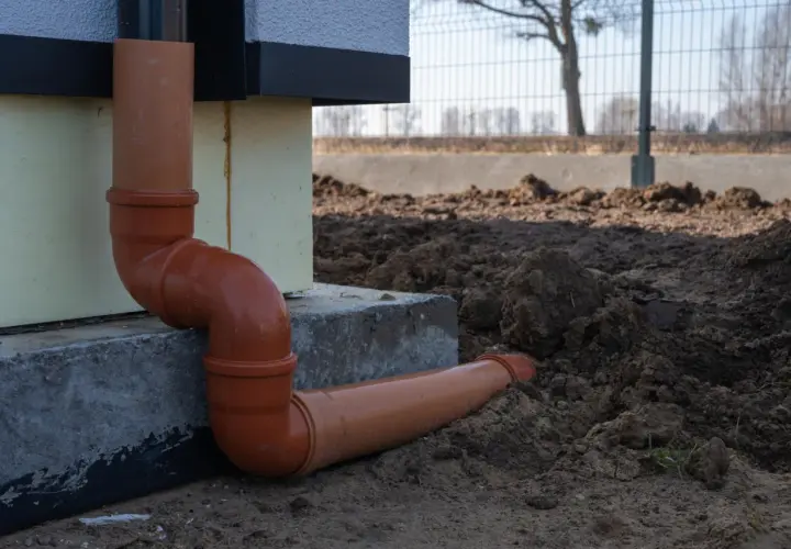 Pomarańczowe rury do odprowadzenia wody z rynien, połączone kolankami, biegną wzdłuż fundamentu domu i wkopane są w ziemię.
