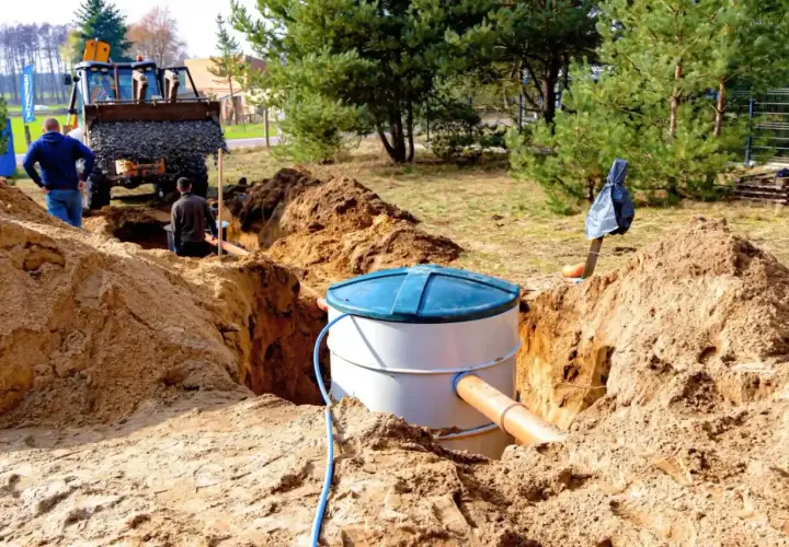 Trwa montaż oczyszczalni przydomowej. Koparka zasypuje wykop, a pracownicy nadzorują instalację zbiornika.
