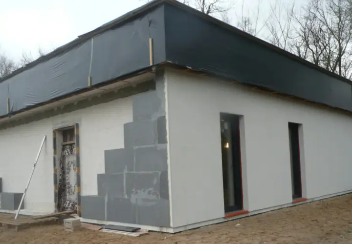 Budowa domu ze styropianu w toku. Ściany z bloczków, częściowo otynkowane, dach przykryty czarną folią.