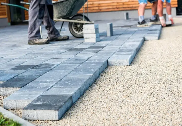 Układanie płyt betonowych na podjeździe – alternatywa dla kostki. Pracownicy budowlani układają szare płyty na podsypce żwirowej.