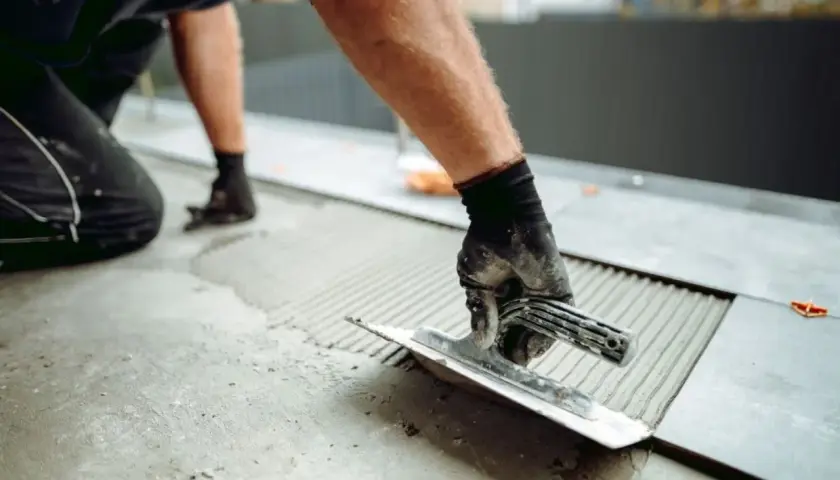 Układanie płytek 60x60 wymaga odpowiedniego kleju. Ręka w rękawicy rozprowadza zaprawę kielnią na podłożu.