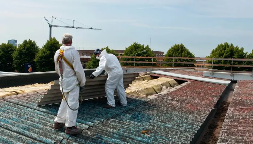 Dwóch pracowników w białych kombinezonach usuwa eternit z dachu. Jeden z nich trzyma duży, pofalowany arkusz.