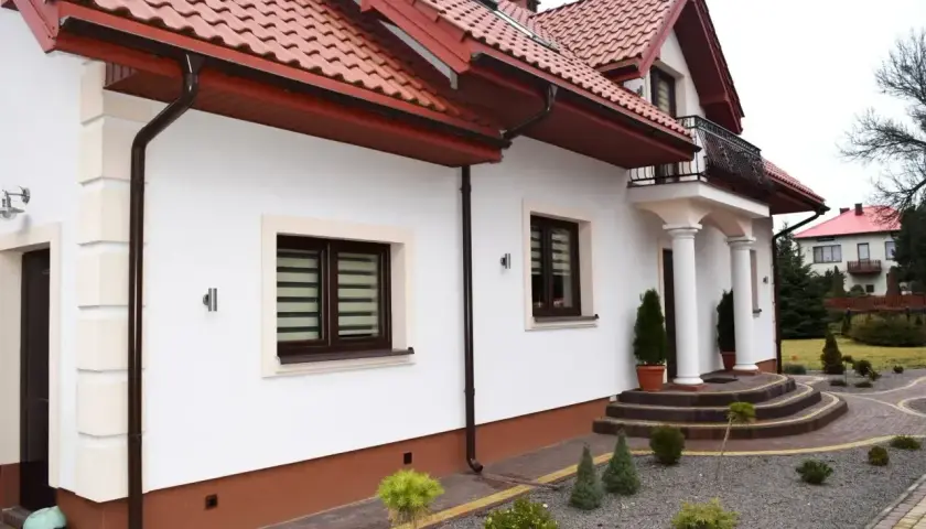 Biały dom z brązowym dachem i cokołem. Eleganckie schody prowadzą do wejścia.