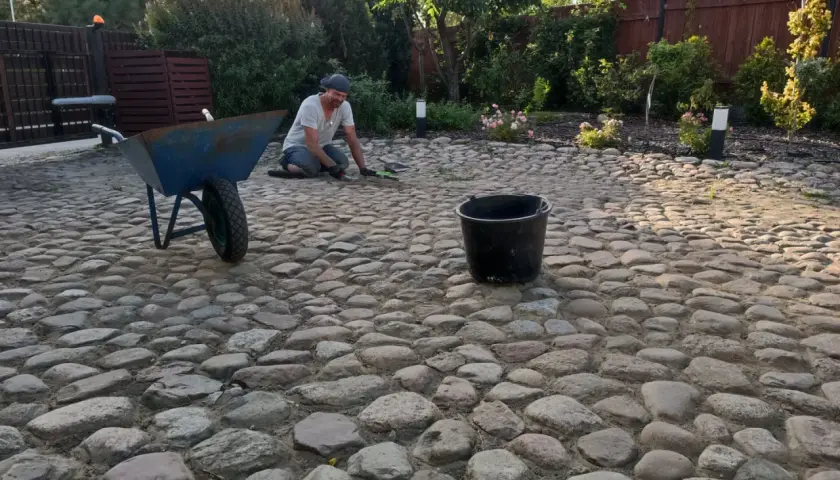 Mężczyzna pracuje przy budowie podjazdu z kamienia polnego. Obok stoi taczka i wiadro.
