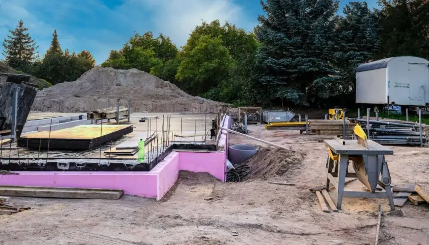 Budowa w stanie zero: fundamenty z różową izolacją, zbrojenie, sterta piasku i sprzęt budowlany.