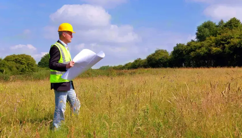 Inżynier w kasku i kamizelce odblaskowej analizuje plany podczas badań geotechnicznych gruntu na łące.