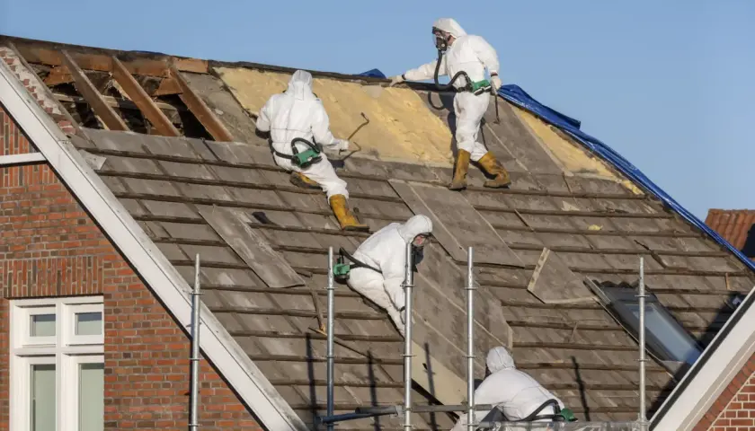 Pracownicy w kombinezonach ochronnych usuwają stare pokrycie dachowe. To praca z materiałami zawierającymi azbest, co to jest, jest niebezpieczne.