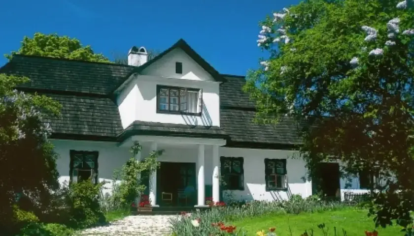 Piękny biały dom z ciemnym dachem, otoczony zielenią i kwitnącymi kwiatami. Widać ścieżkę z kamieni i ganek, jakby zrealizowano dobudowa ganku projekt.
