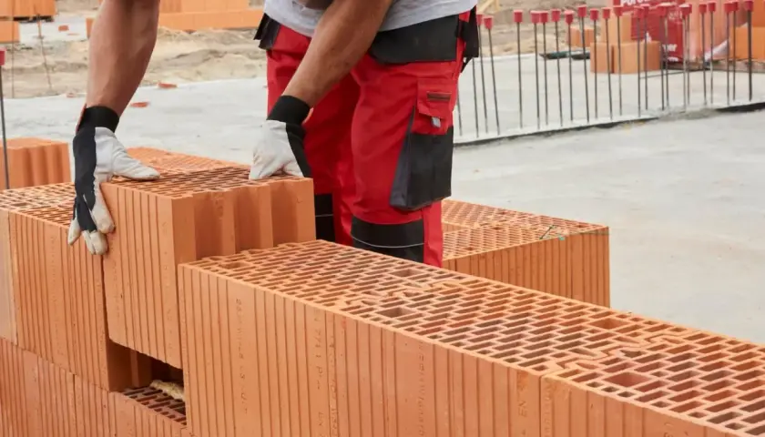 Budowa domu z pustaków ceramicznych Porotherm. Pracownik w czerwonych spodniach i rękawicach układa kolejne elementy.