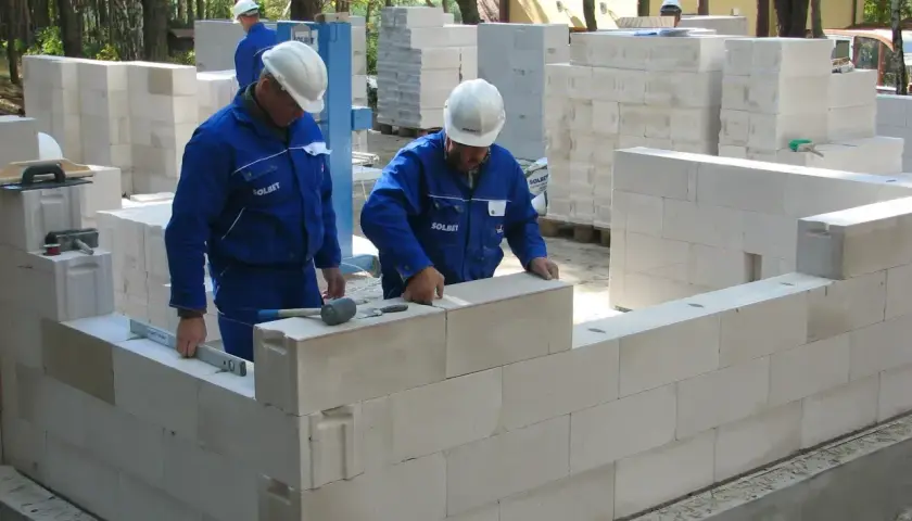 Budowa domu z bloczków. Pracownicy w niebieskich kombinezonach układają ściany. Wybór materiału: solbet czy ytong?