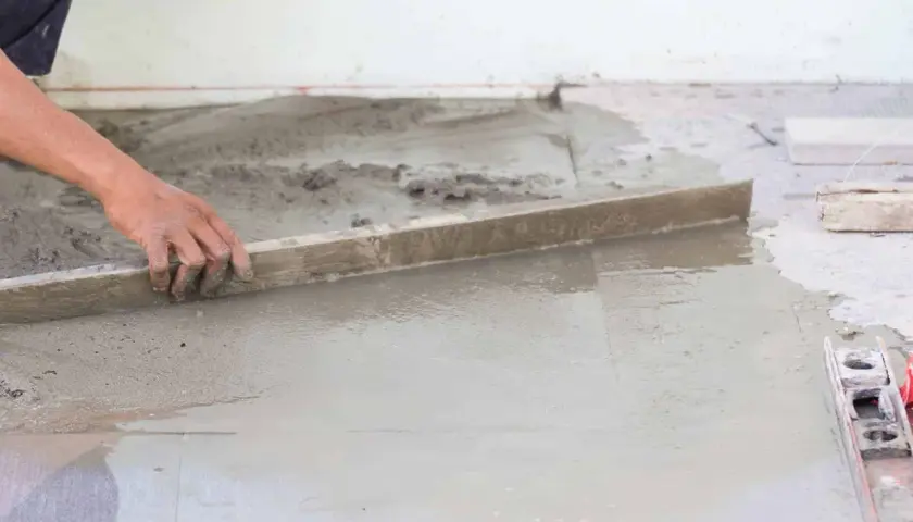 Ręka wyrównuje świeżą posadzkę na gruncie za pomocą łaty.