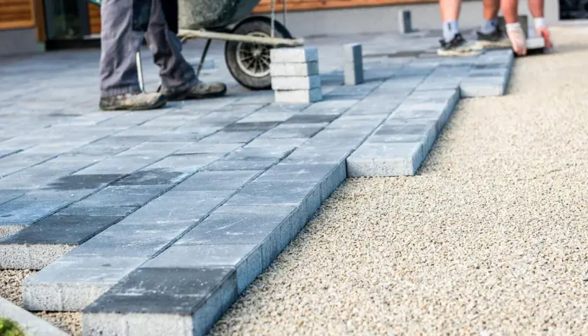 Układanie płyt betonowych na podjeździe – alternatywa dla kostki. Pracownicy budowlani układają szare płyty na podsypce żwirowej.