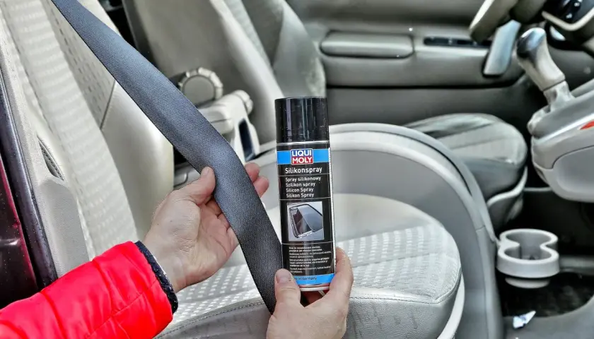 Ręce trzymające pas bezpieczeństwa i puszkę z napisem "Silikonspray". Preparat ten to olej silikonowy do paneli, który ułatwia konserwację wnętrza samochodu.