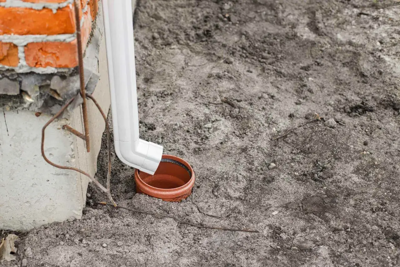 Biała rura spustowa odprowadza wodę z rynien do brązowej studzienki.
